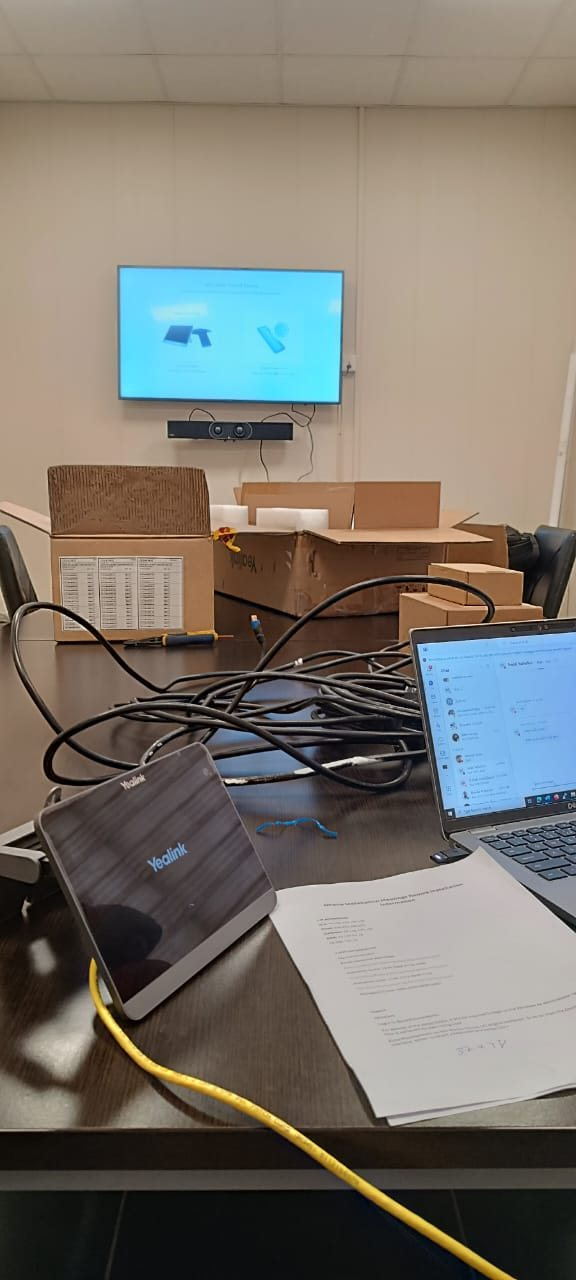 A meeting room part-way through an audio-visual installation: a wall-mounted display and soundbar, unpacked Yealink room kit, cabling and an engineer’s laptop showing the build sheet.