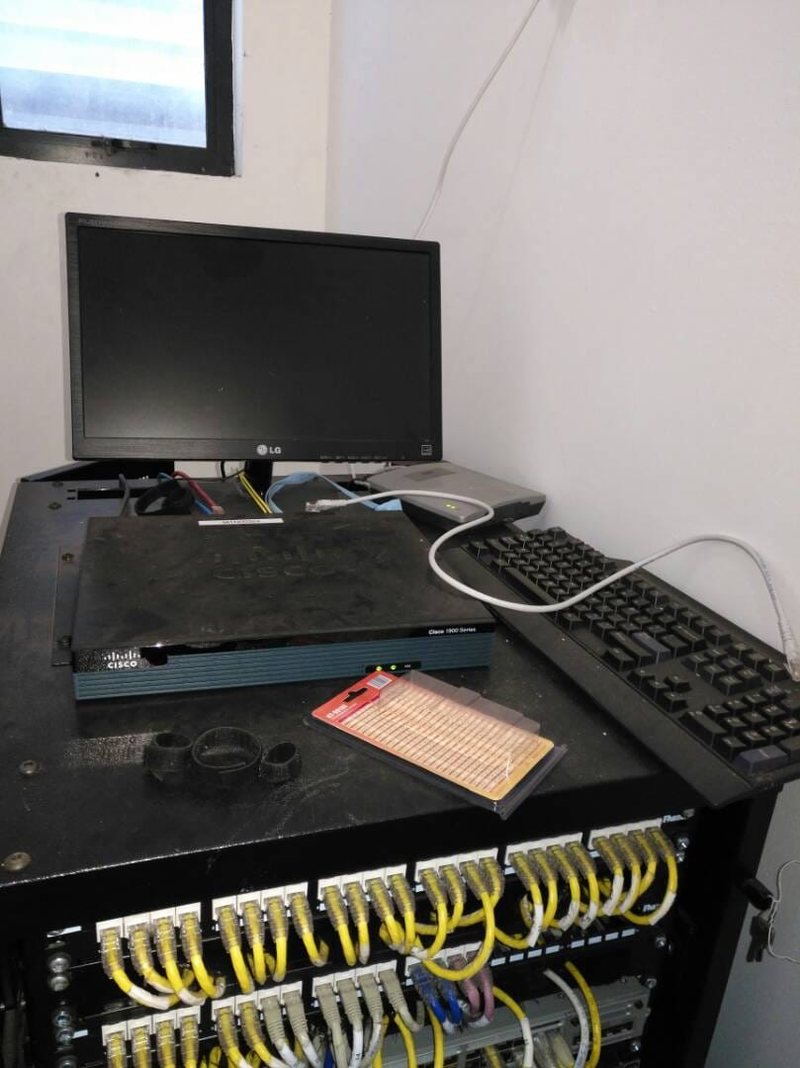 A comms room cabinet with a Cisco 1900 Series router, patch panel and yellow structured cabling, with a monitor and keyboard on top for console access.