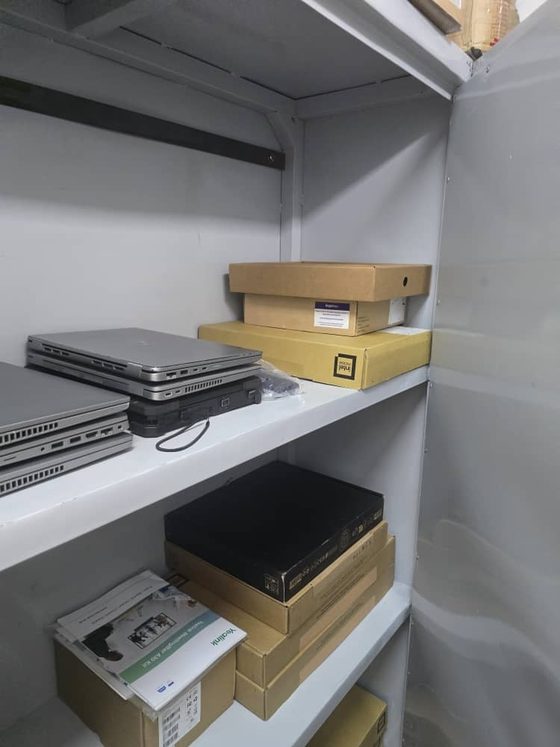 Matériel informatique préparé sur des rayonnages d'entrepôt : ordinateurs portables empilés, un poste de travail compact, un kit de salle de visioconférence en carton et des pièces de rechange conservés pour des interventions planifiées.